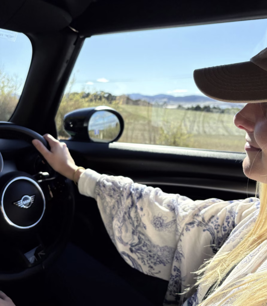 A woman driving a Mini Cooper. Half a steering wheel, her profile, cap, and arm. Out the window is the Tasmanian landscape.
